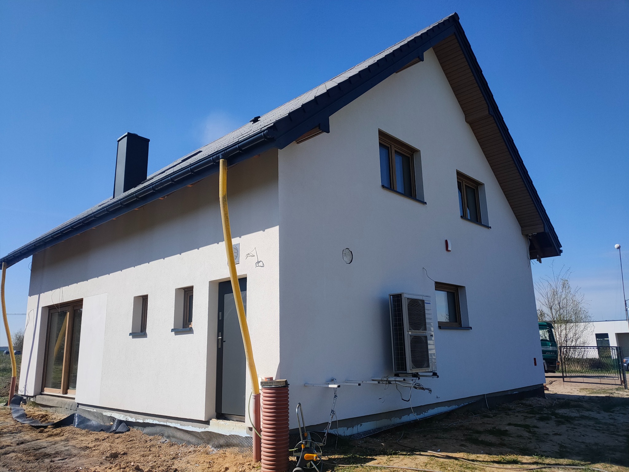 Nowoczesny dom jednorodzinny z białą elewacją i ciemną dachówką, widoczny z zewnątrz w słoneczny dzień, z zamontowanym klimatyzatorem na ścianie i żółtą rurą spustową.