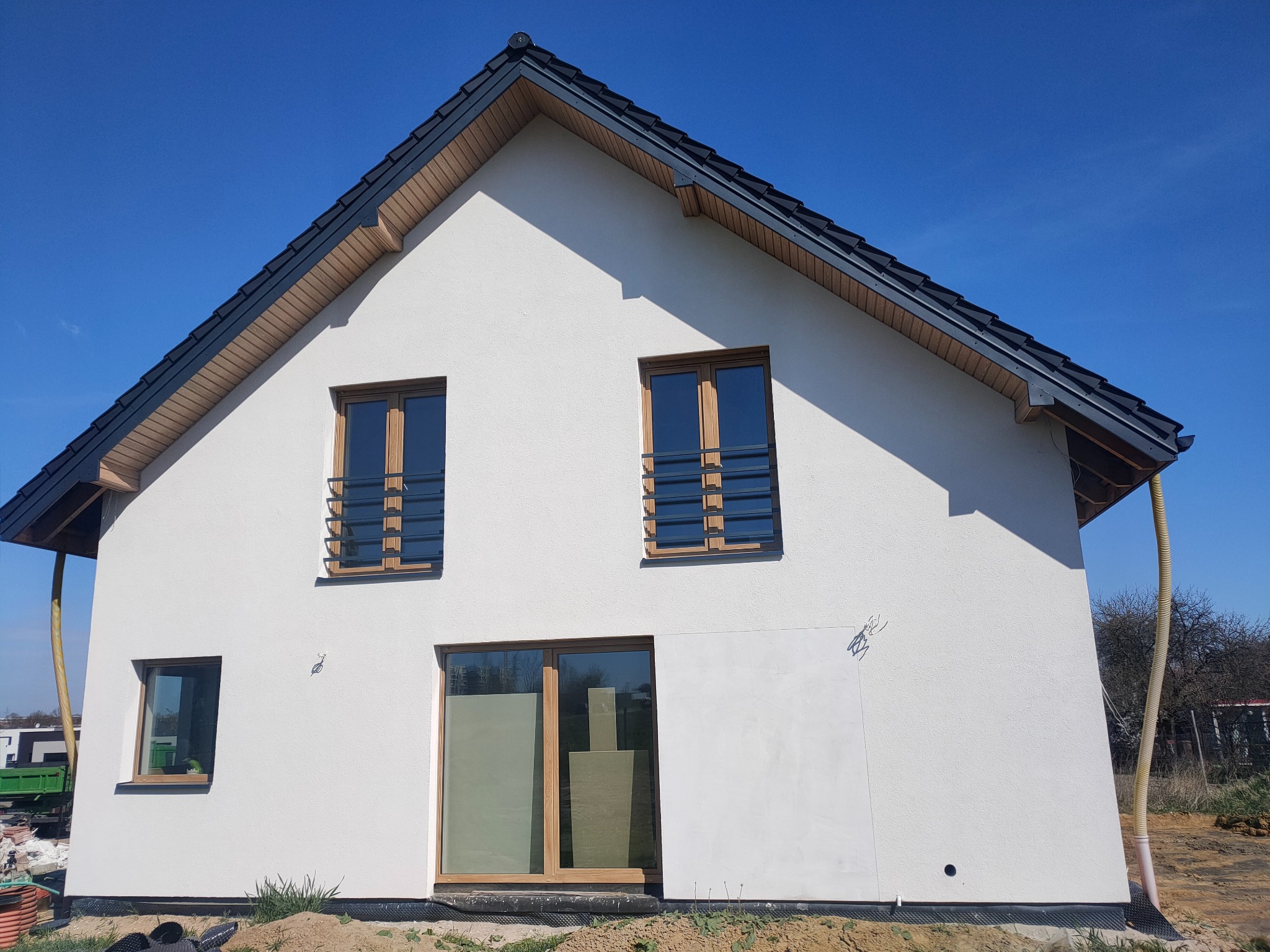 Nowo otynkowany dom jednorodzinny z ciemną dachówką i drewnianą stolarką okienną, widoczne rury wentylacyjne przy elewacji bocznej, teren wokół domu jeszcze niezagospodarowany.