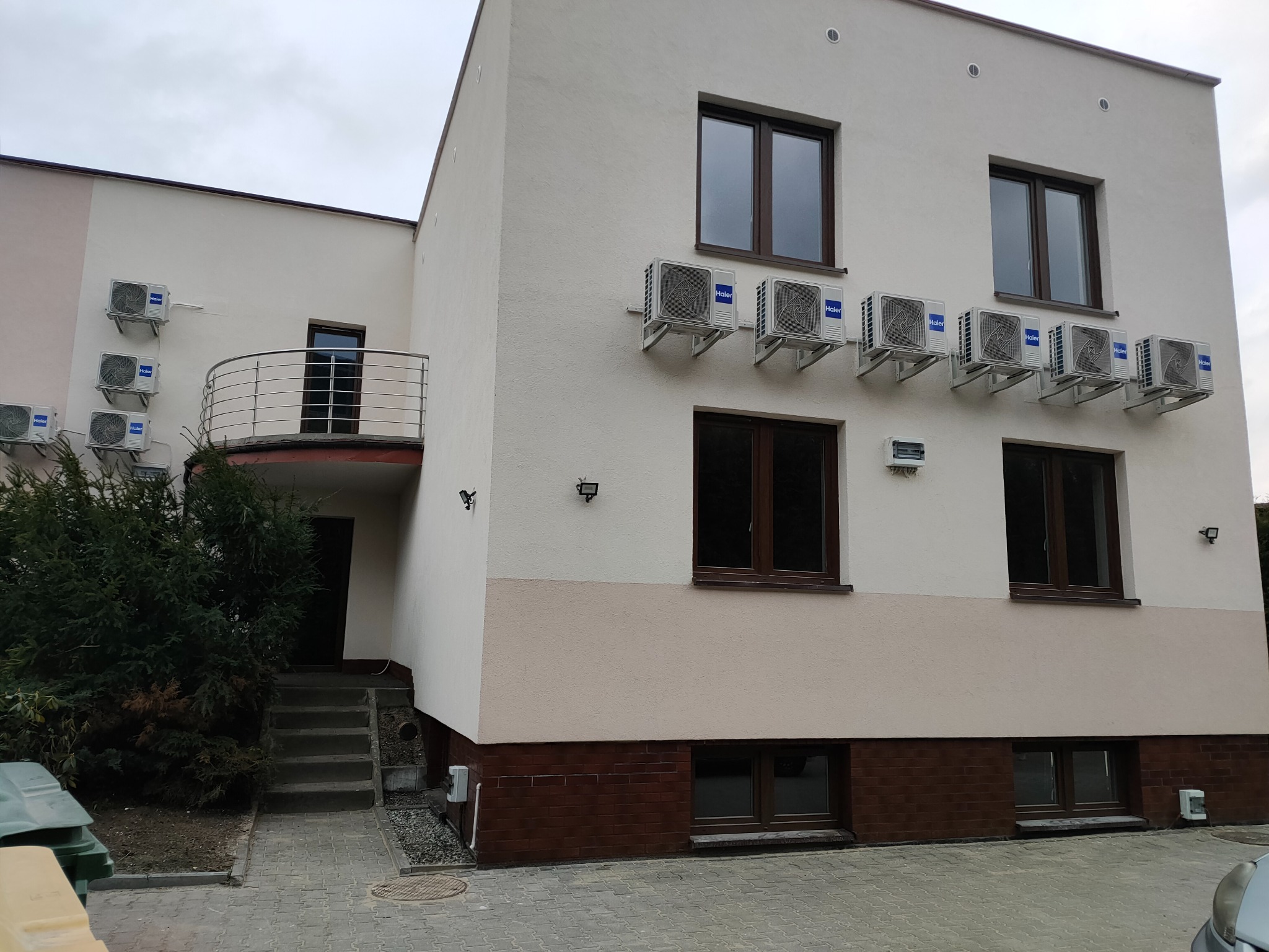 Dwukondygnacyjny budynek z jasną elewacją, ciemnymi oknami i rzędem klimatyzatorów na ścianie. Widoczne schody wejściowe i niewielki balkon z metalową barierką.