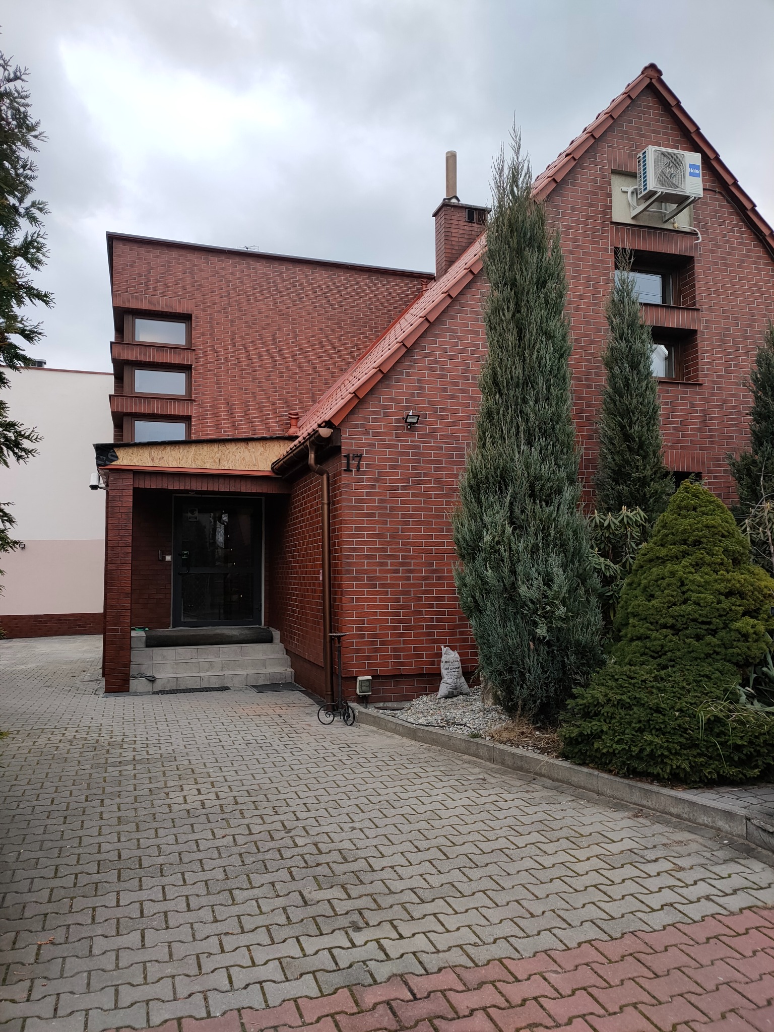 Ceglany dom jednorodzinny z numerem 17, widoczne elementy elewacji, okna, dach, klimatyzator, roślinność i chodnik z kostki. Architektura z elementami nowoczesnymi.