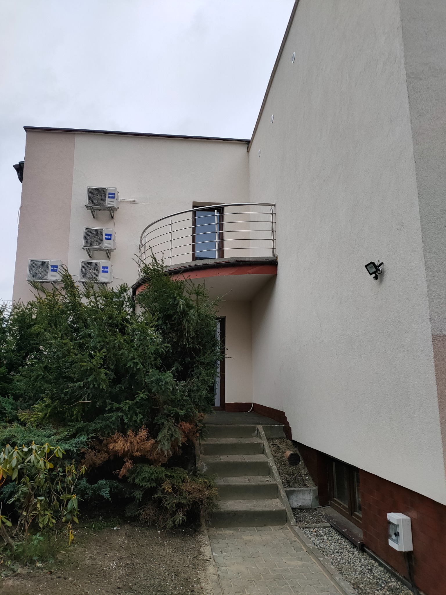 Dwukondygnacyjny budynek z jasną elewacją, balkonem z metalową balustradą i trzema klimatyzatorami na ścianie. Widoczne schody wejściowe i oświetlenie zewnętrzne.