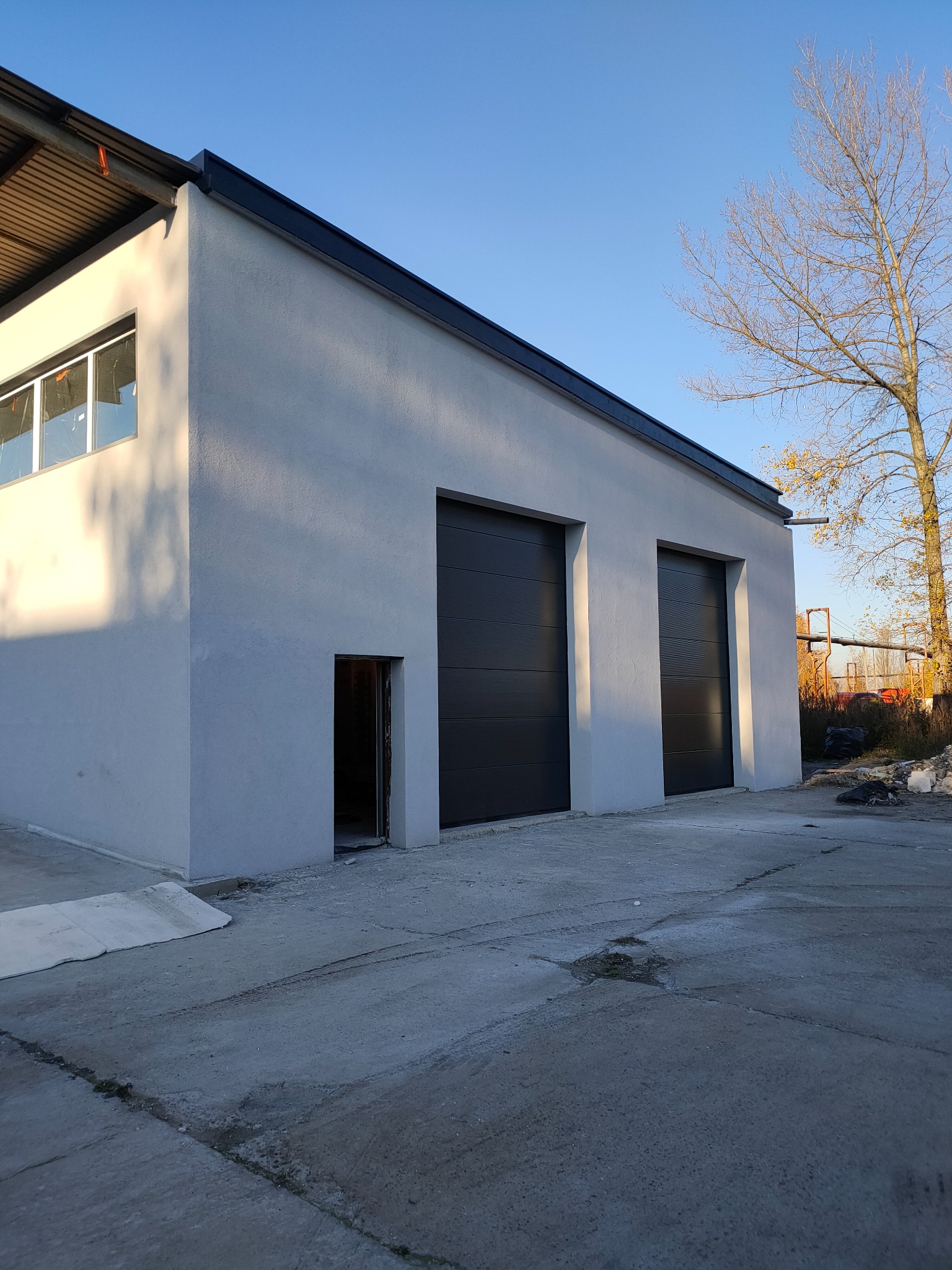 Nowoczesny budynek z szarą elewacją i dwoma czarnymi bramami garażowymi w słoneczny dzień. Widoczne okno i otwarte wejście. Minimalistyczny design.