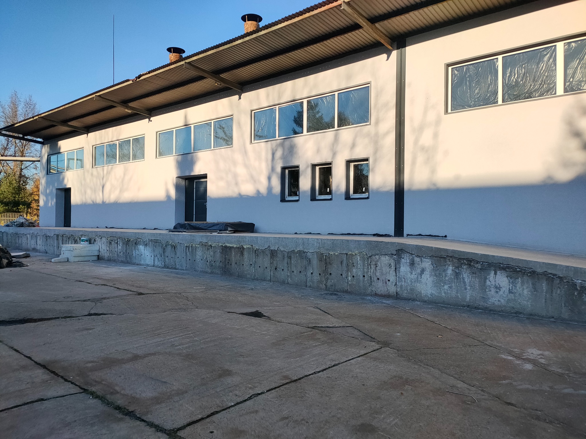 Szary budynek przemysłowy po renowacji elewacji, z nowymi oknami zabezpieczonymi folią. Widoczny betonowy podjazd i cień drzewa na ścianie.