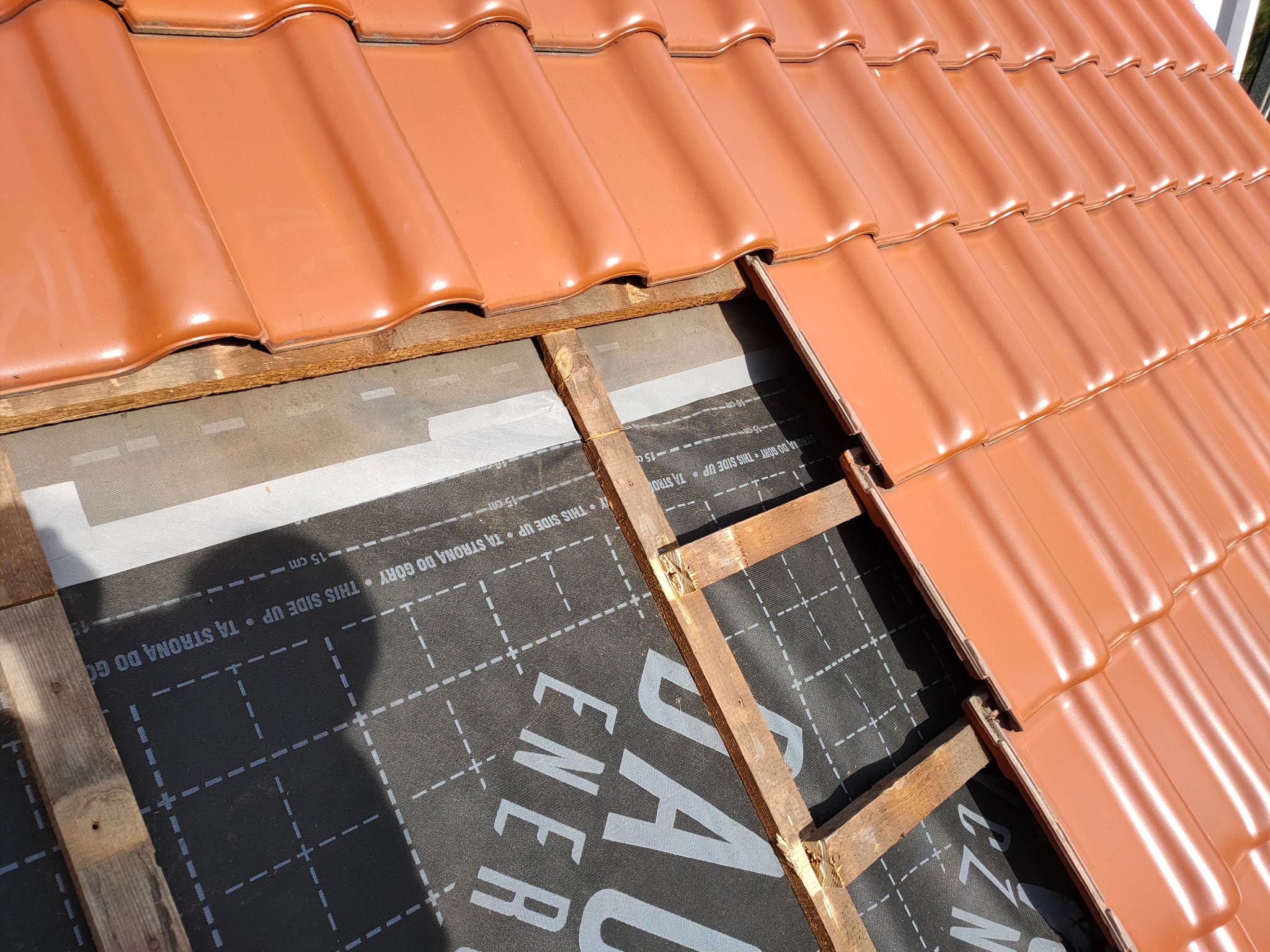 Fragment dachu z częściowo ułożoną czerwoną dachówką, widoczna membrana dachowa z nadrukiem. Elementy drewniane konstrukcji dachu. Ujęcie z góry, w słoneczny dzień.