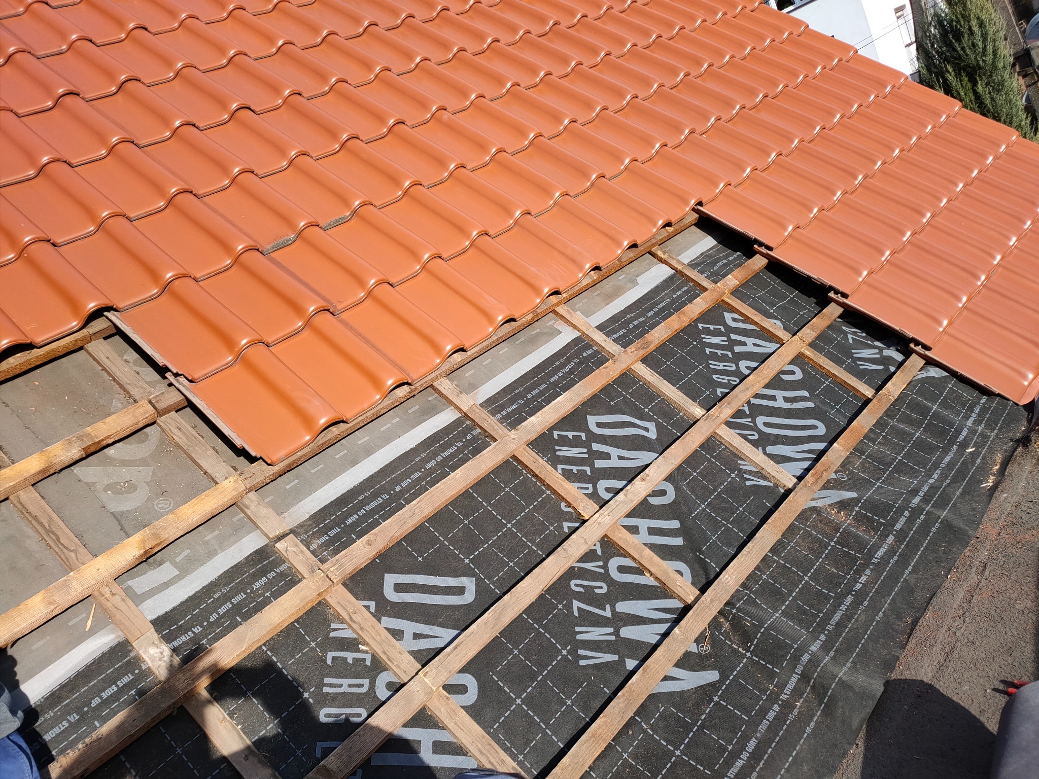 Częściowo ułożona, nowa, pomarańczowa dachówka ceramiczna na łatach i membranie dachowej. Widoczny fragment budynku w tle. Słoneczny dzień.