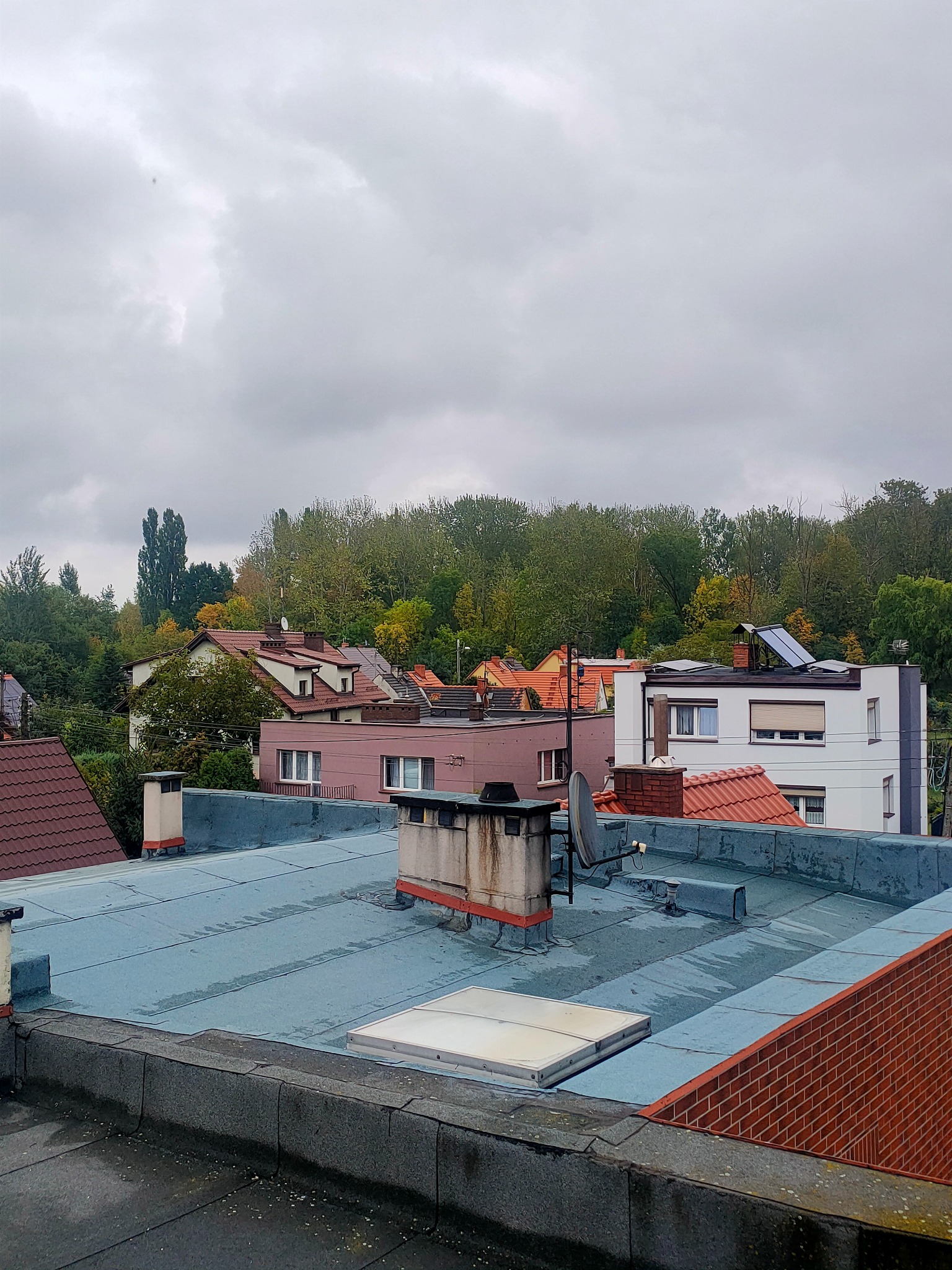 Widok na dachy budynków z płaskiego dachu pokrytego papą, kominy i anteny satelitarne na tle drzew i pochmurnego nieba. Widoczne panele słoneczne na jednym z budynków.