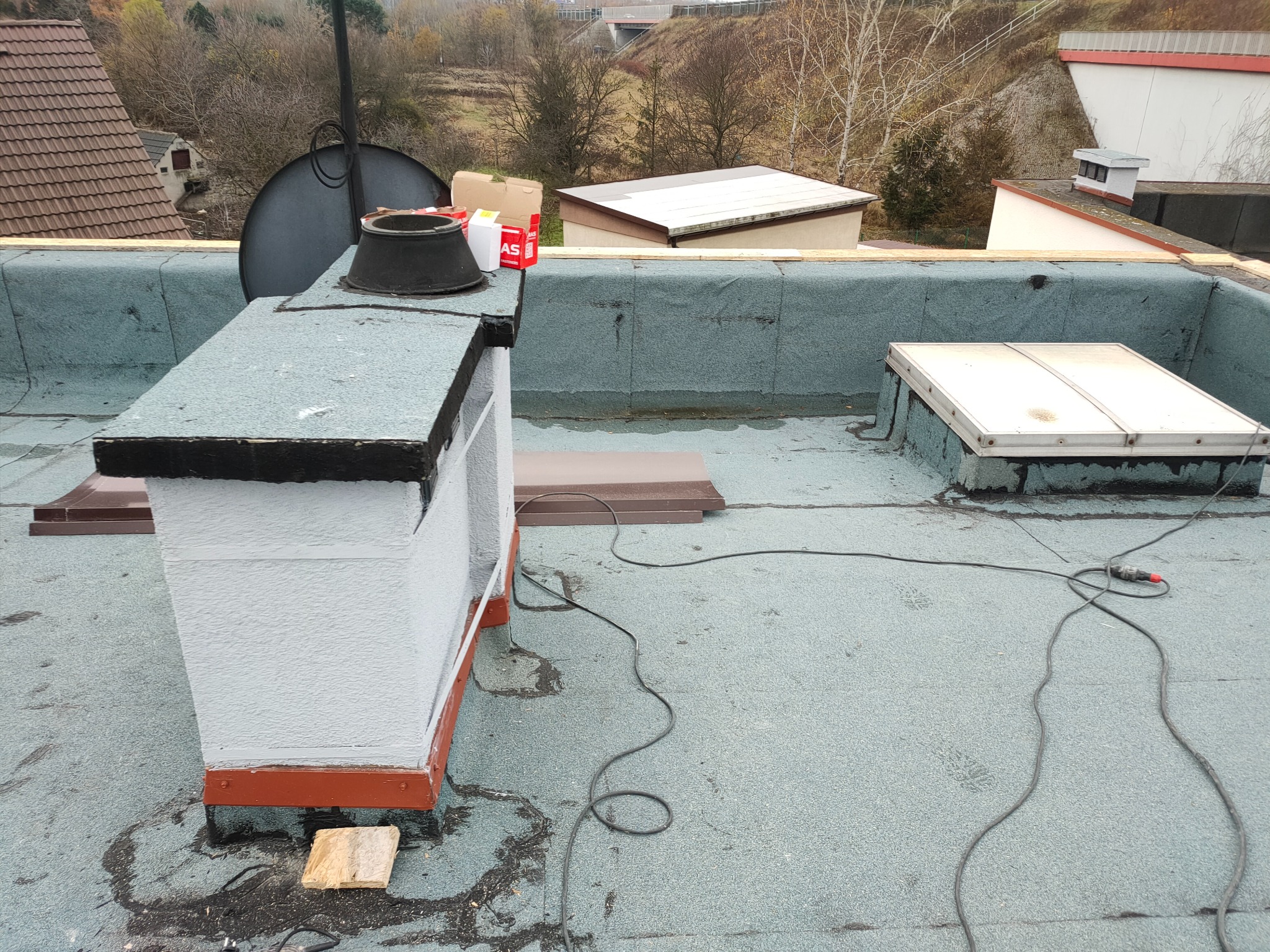 Płaski dach pokryty papą z kominem w trakcie renowacji. Widoczne narzędzia i materiały budowlane. W tle zabudowania i drzewa.