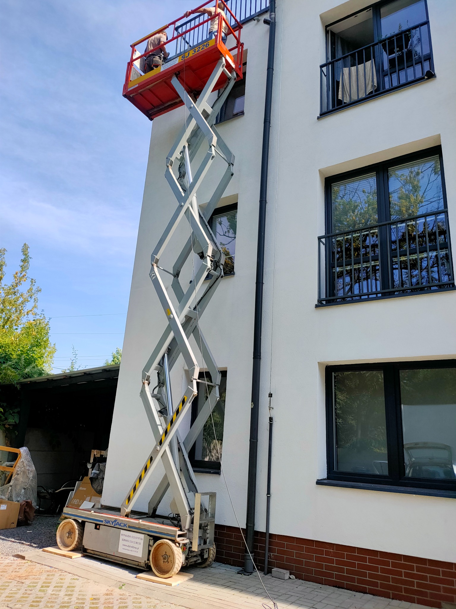 Praca na wysokości: Dwóch pracowników na podnośniku koszowym maluje białą elewację budynku z czarnymi oknami i balkonami. Widok z dołu, słoneczny dzień.