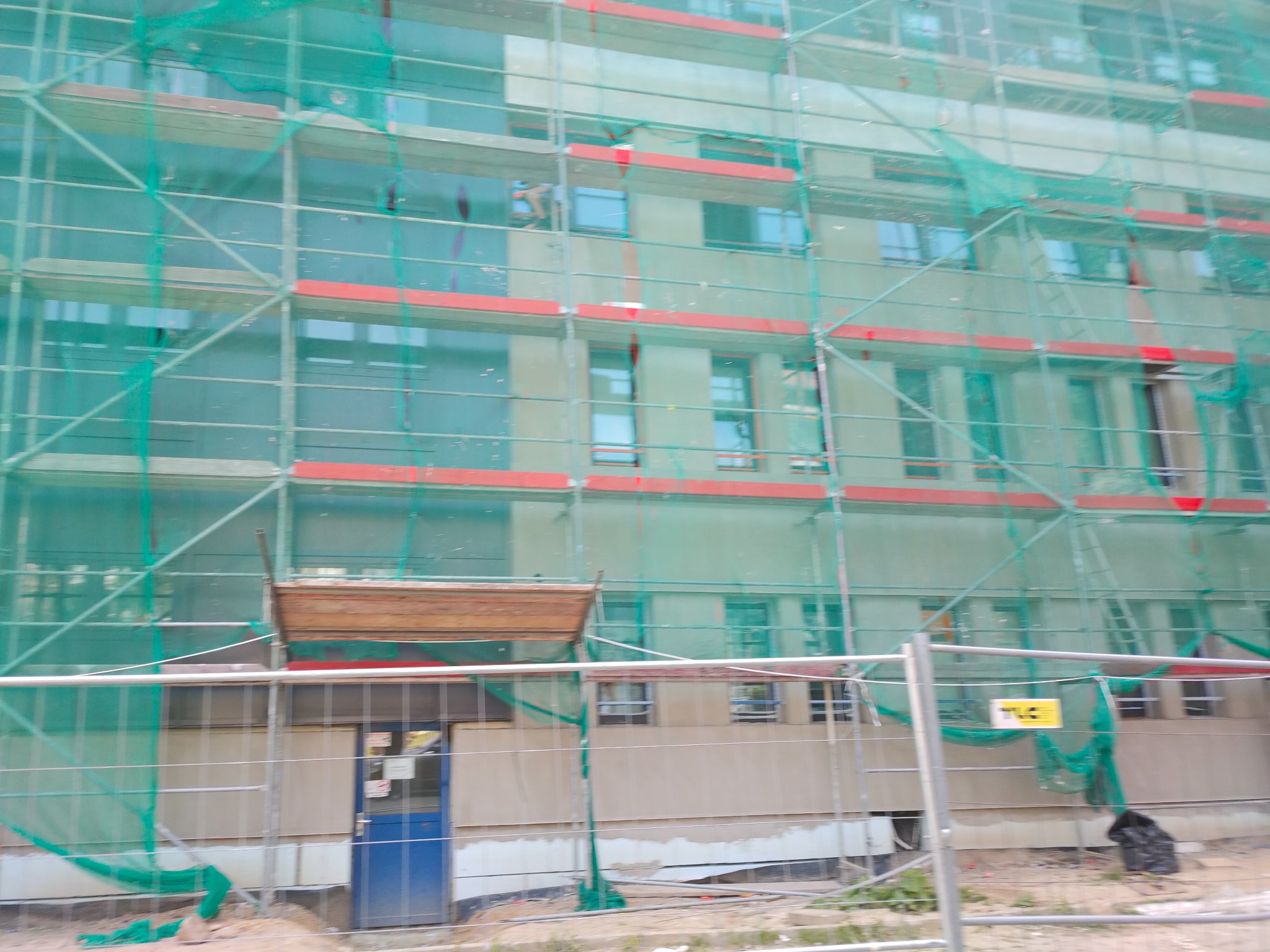 Budynek w trakcie prac elewacyjnych, osłonięty siatką ochronną i rusztowaniami. Widoczne okna i fragmenty elewacji. Ujęcie z dołu, zza ogrodzenia.