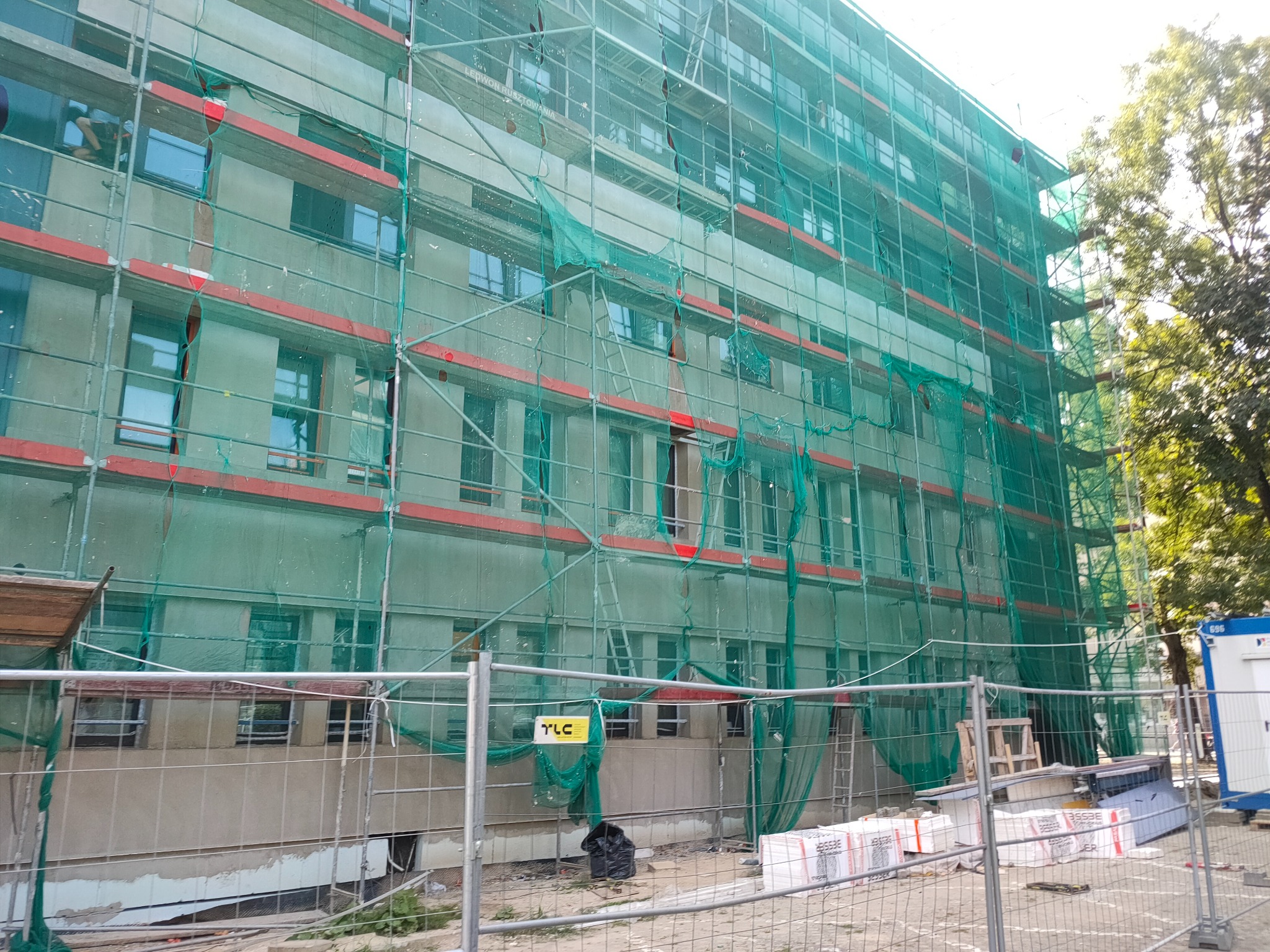 Budynek w trakcie prac elewacyjnych, osłonięty siatką rusztowaniową. Widoczne rusztowania, materiały budowlane i fragment zieleni. Ujęcie z dołu, w dzień.