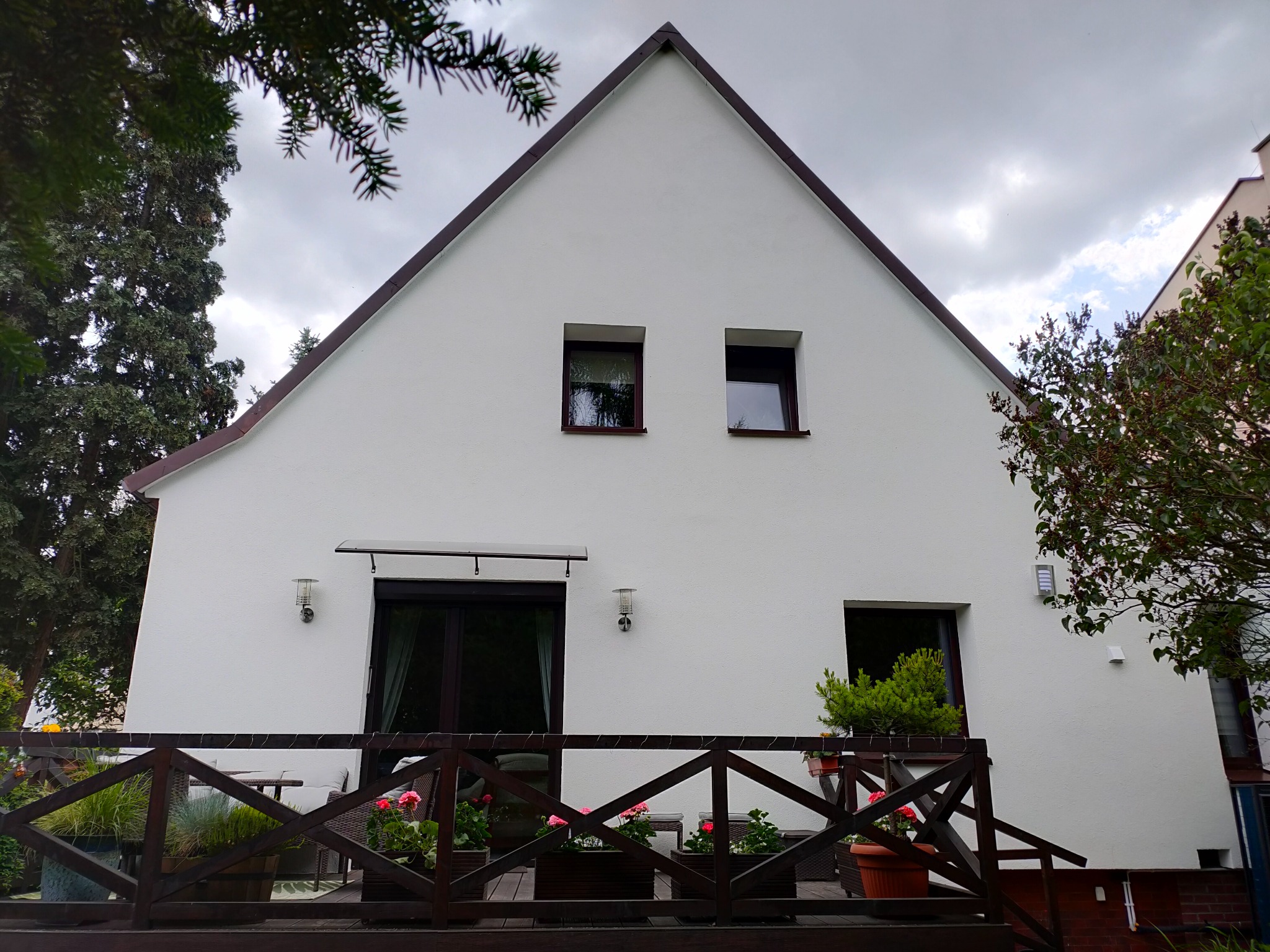 Biały dom z brązowym dachem widziany z tarasu z drewnianą balustradą i kwiatami w donicach. Widoczne okna i drzwi wejściowe. Pochmurne niebo w tle.