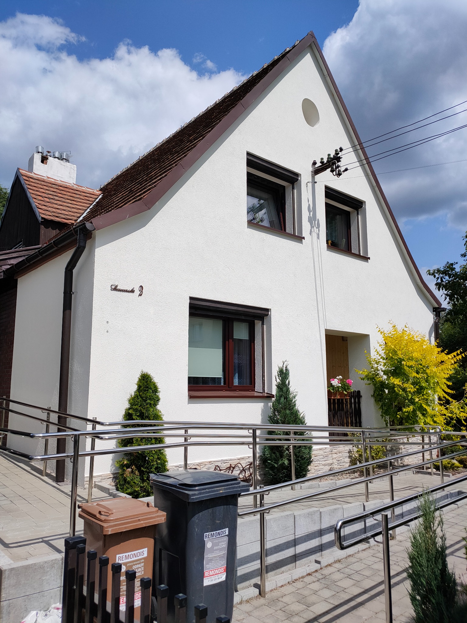 Biały dom z brązowym dachem i oknami, widoczny z zewnątrz, z rampą dla niepełnosprawnych i dwoma koszami na śmieci przed wejściem. Słoneczny dzień, błękitne niebo z chmurami.