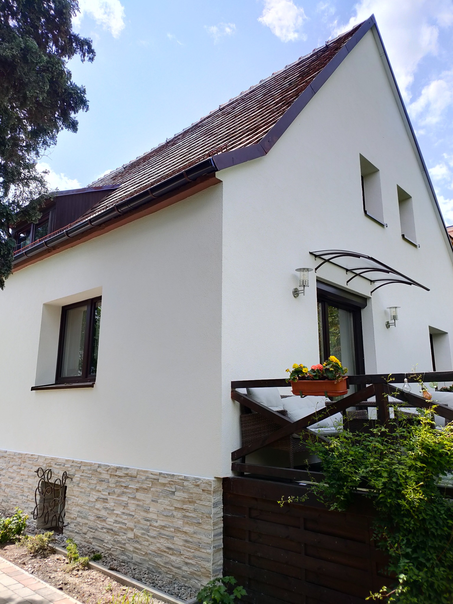 Biały dom z brązowym dachem i elementami dekoracyjnymi z kamienia. Widoczny balkon z kwiatami i zadaszenie nad drzwiami. Elewacja po renowacji w słoneczny dzień.