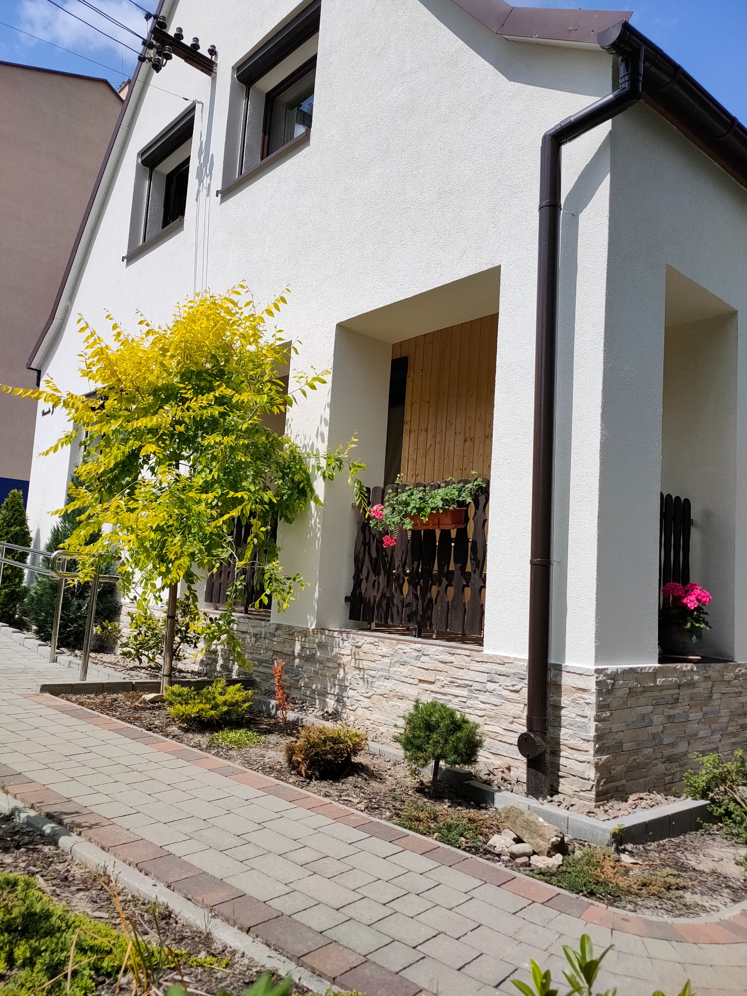 Biała elewacja domu z dekoracyjnym kamieniem, brązowymi rynnami i stolarką okienną. Żółte drzewko przy ścianie. Ujęcie z poziomu chodnika.