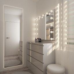 KORANTI Izabela Kornacka - Jasna toaletka z lustrem podświetlanym żarówkami, biała komoda z szufladami i pufa w minimalistycznym, nowoczesnym wnętrzu. Widoczne kosmetyki i fragment okna.