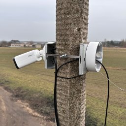 Sławomir Zapora FIREWALL - Kamera monitoringu Dahua zamontowana na betonowym słupie z widoczną puszką montażową, kablem i metalową obejmą, na tle pola i zabudowań w oddali.