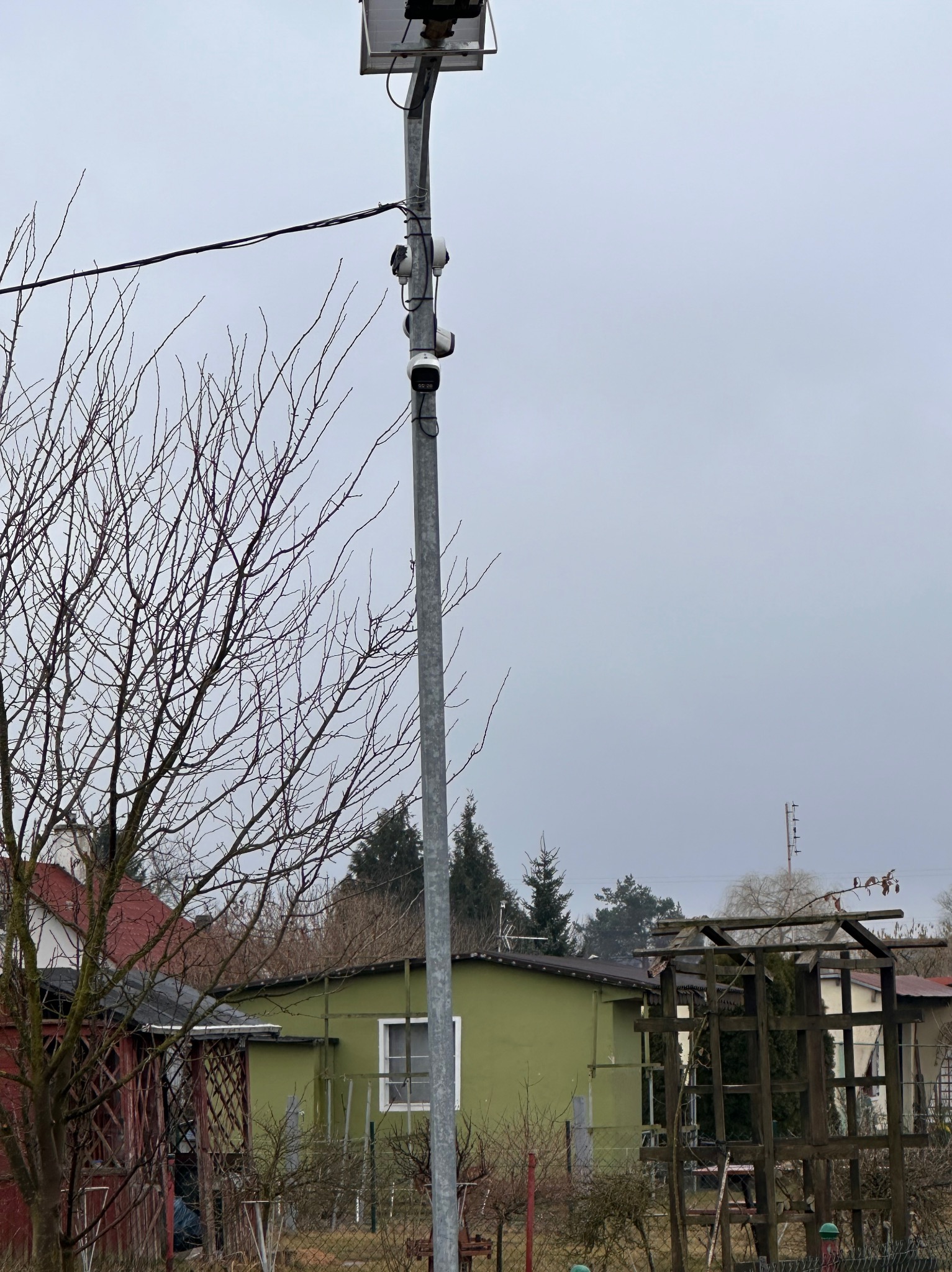 Słup z panelem solarnym i kilkoma kamerami monitoringu zainstalowany na zewnątrz, na tle pochmurnego nieba i zabudowań działkowych.