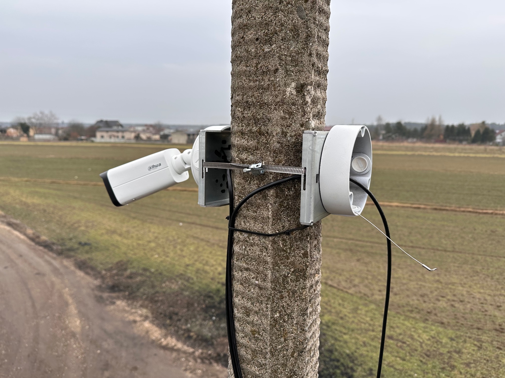 Kamera monitoringu Dahua zamontowana na betonowym słupie z widoczną puszką montażową, kablem i metalową obejmą, na tle pola i zabudowań w oddali.