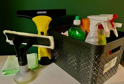 Zestaw narzędzi do czyszczenia okien: elektryczna myjka Karcher, spryskiwacz z nakładką z mikrofibry, ściereczki i pojemnik z detergentami na białym blacie, na tle ciemnozielonej ściany.