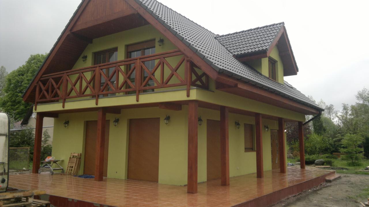 Dwukondygnacyjny budynek z balkonem z drewnianą balustradą w kształcie krzyży, zadaszony taras wyłożony płytkami, elewacja w odcieniach zieleni, szara dachówka, widoczne oświetlenie zewnętrzne,...