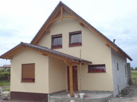 Niedokończony budynek jednorodzinny z drewnianą konstrukcją dachu i werandy, częściowo otynkowany, z drewnianymi ramami okiennymi i skrzynkami z kwiatami.