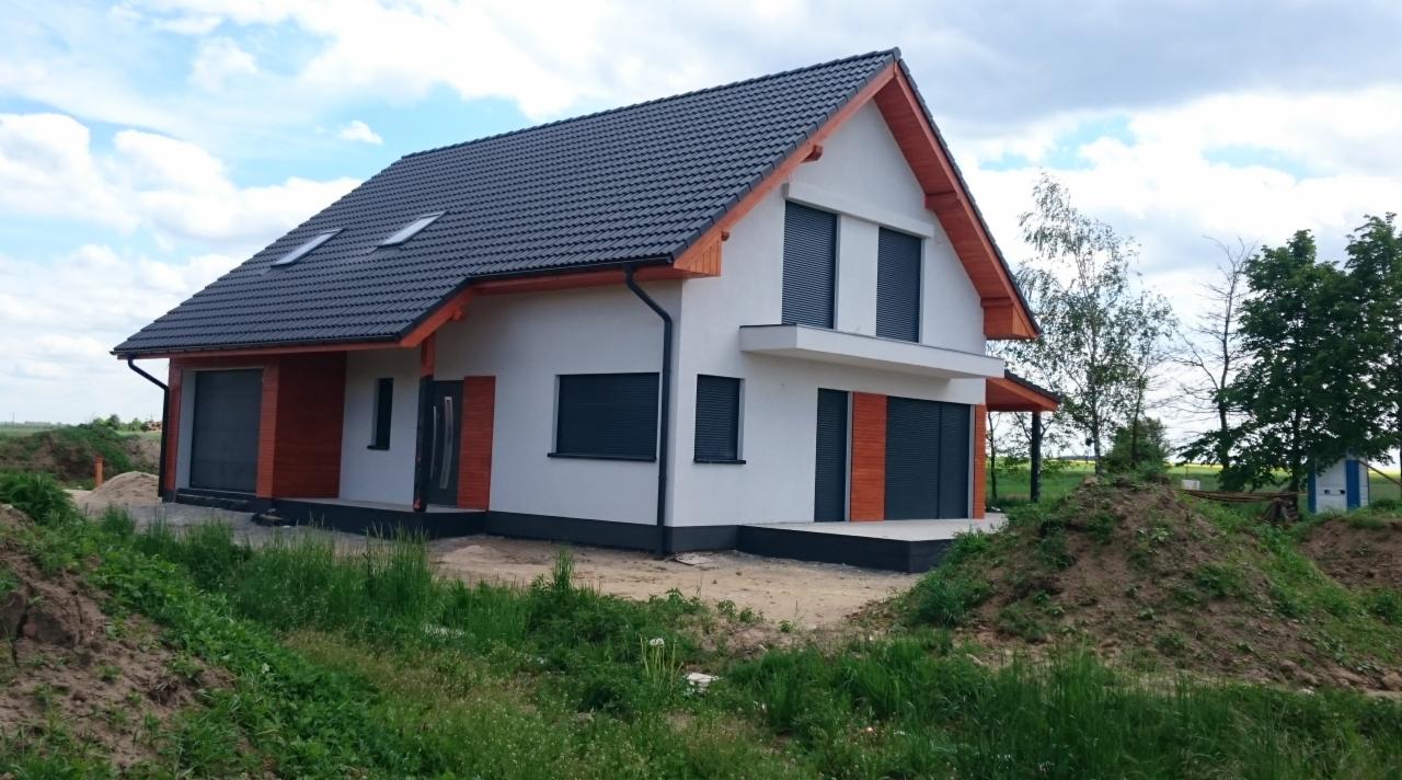 Nowoczesny dom z garażem, wykończony w bieli z elementami drewna w kolorze pomarańczowym, ciemna dachówka, rolety zewnętrzne, trawa na pierwszym planie.