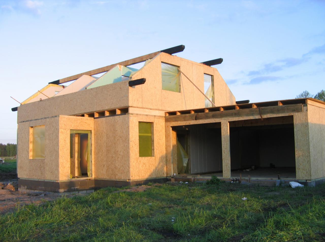 Surowy stan budowy domu z płyty OSB z otwartym garażem, widoczne elementy konstrukcyjne dachu, zielona trawa na pierwszym planie.