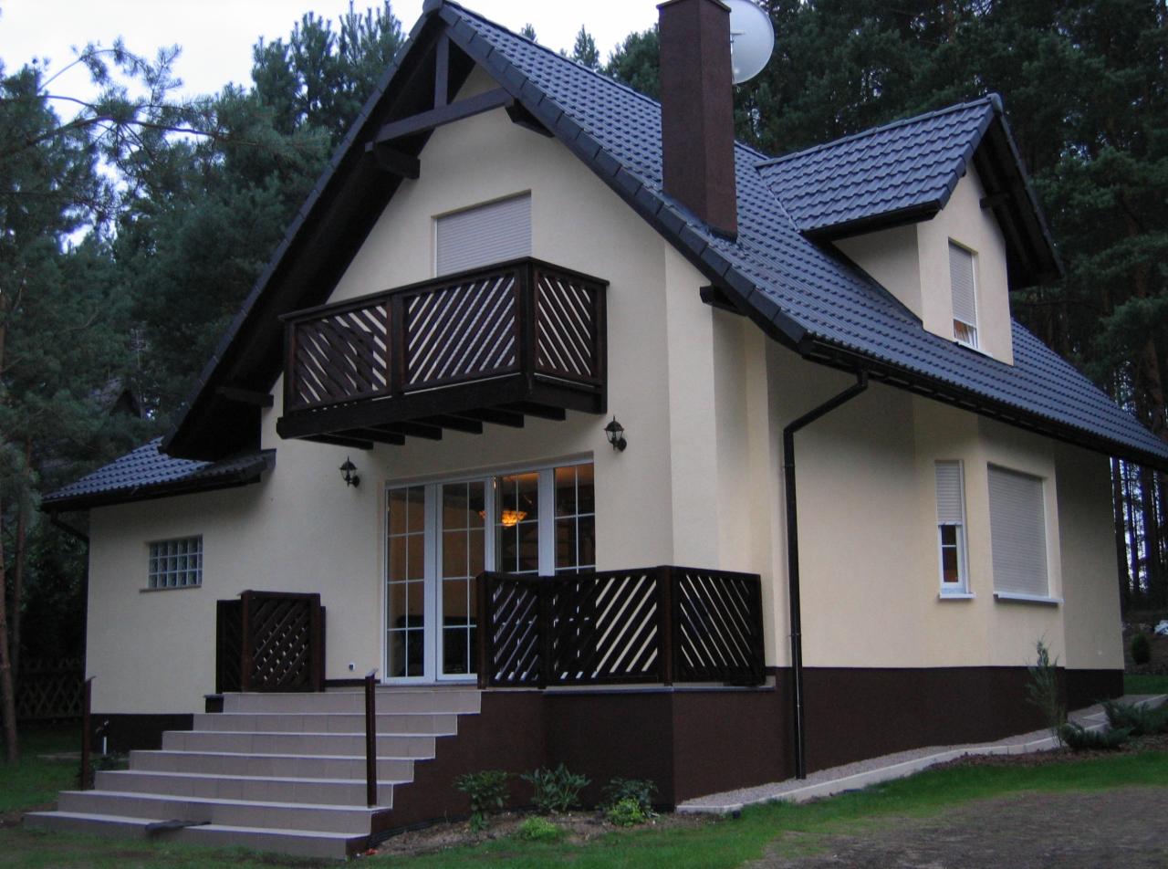 Gotowy dom z balkonem i schodami wejściowymi, ciemna dachówka, kremowa elewacja z brązowym cokołem, na tle lasu.