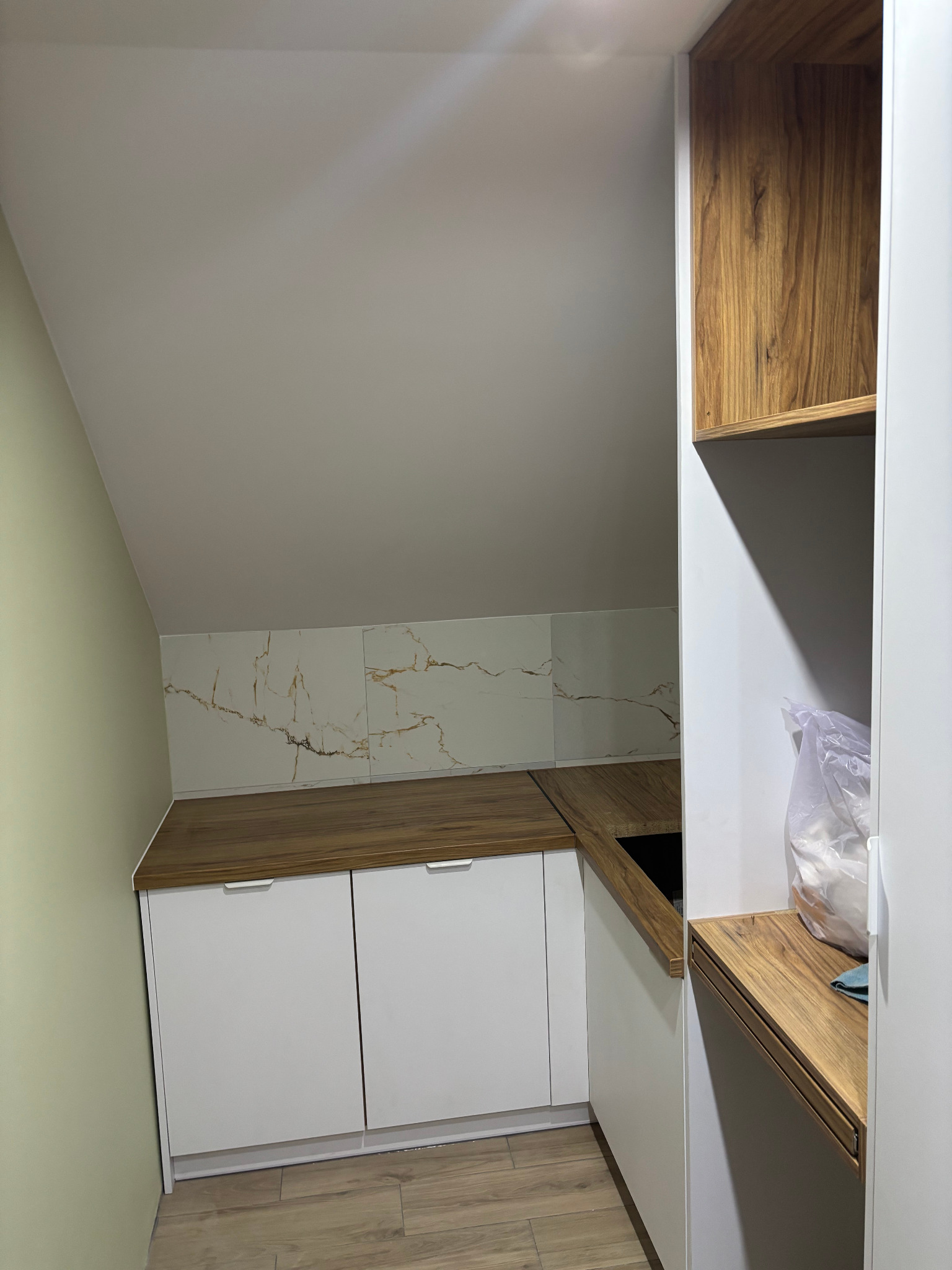 Kuchnia na poddaszu z białymi szafkami, drewnianym blatem i marmurową ścianą nad blatem, widok z dołu na skośny sufit.