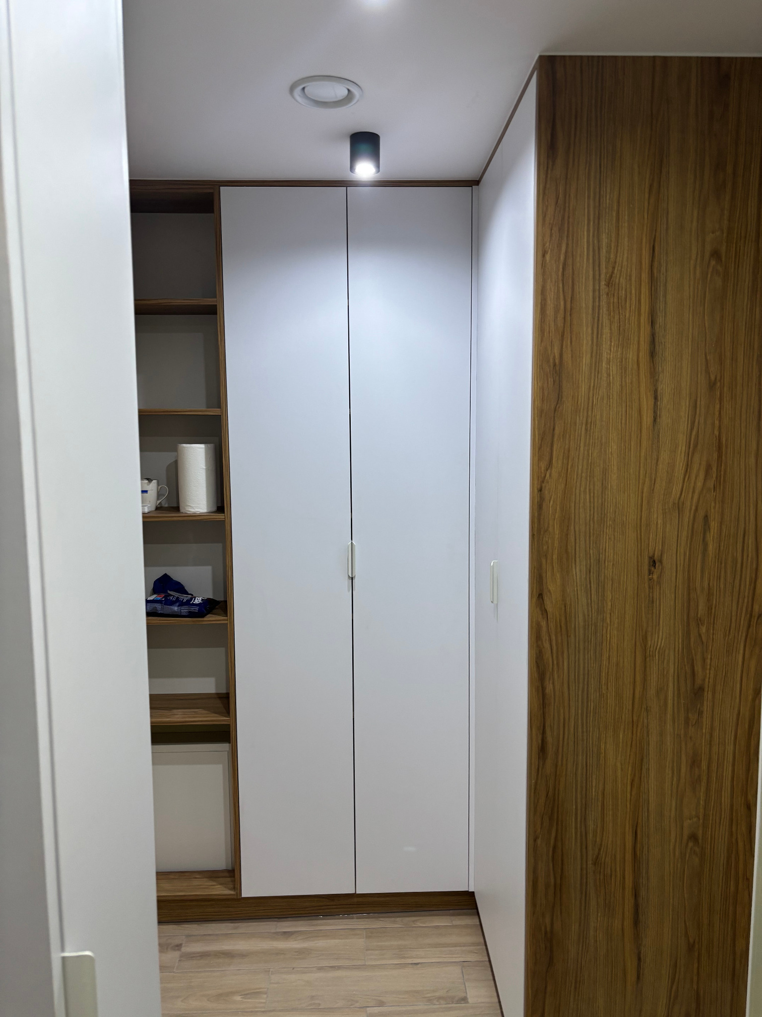 Wnętrze szafy wnękowej z białymi frontami i drewnianymi bokami, widok z wnętrza pomieszczenia, na półkach widoczne przedmioty codziennego użytku.