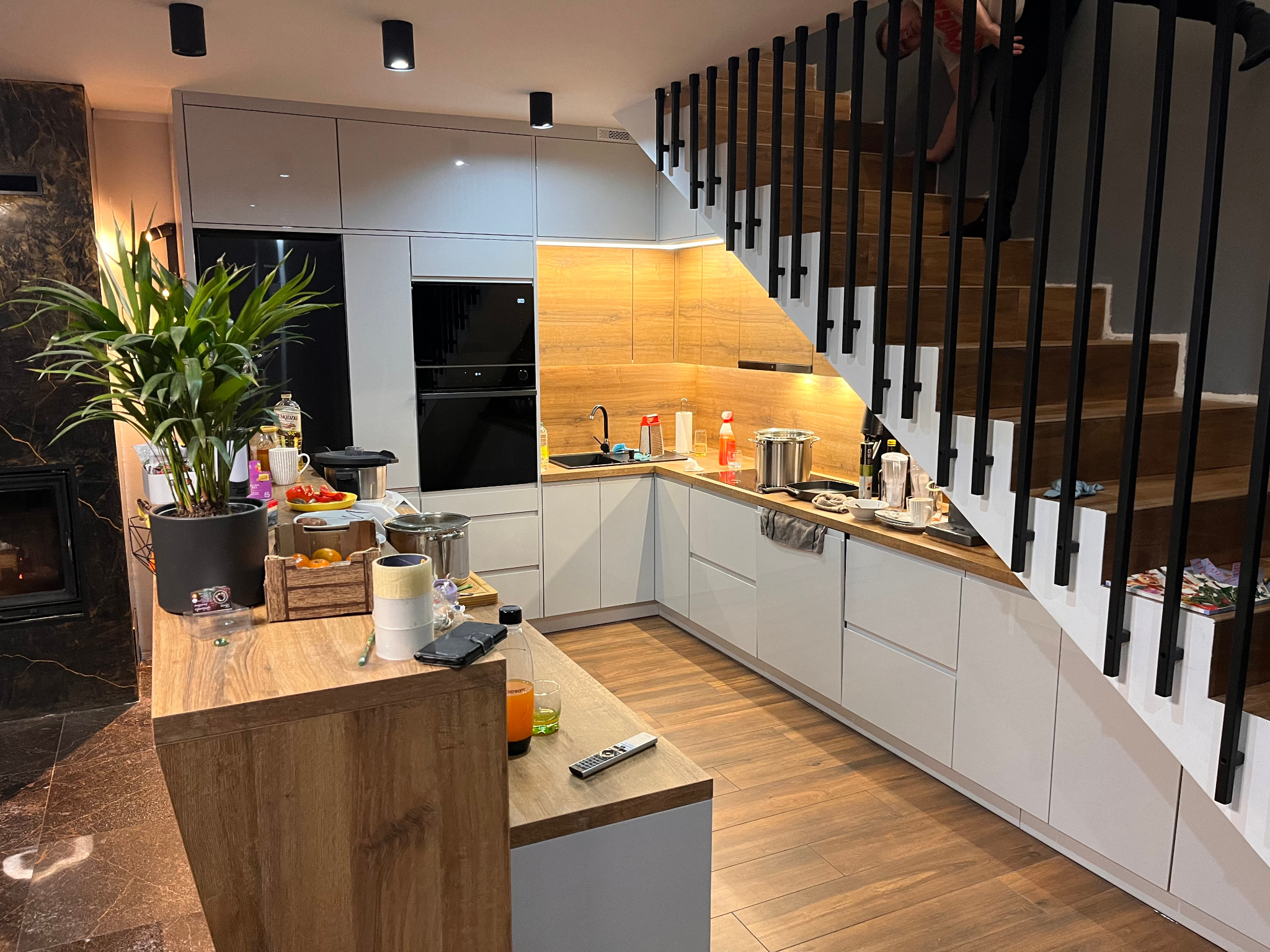 Nowoczesna kuchnia z białymi szafkami i drewnianymi blatami, oświetlona punktowymi lampami, z widocznymi schodami z czarnymi barierkami i drewnianymi stopniami w tle, oraz elementami wyposażenia...