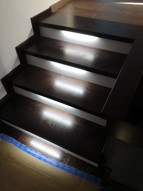 Ciemne, drewniane schody z podświetleniem LED na każdym stopniu, widoczne niebieskie taśmy zabezpieczające na podłodze.