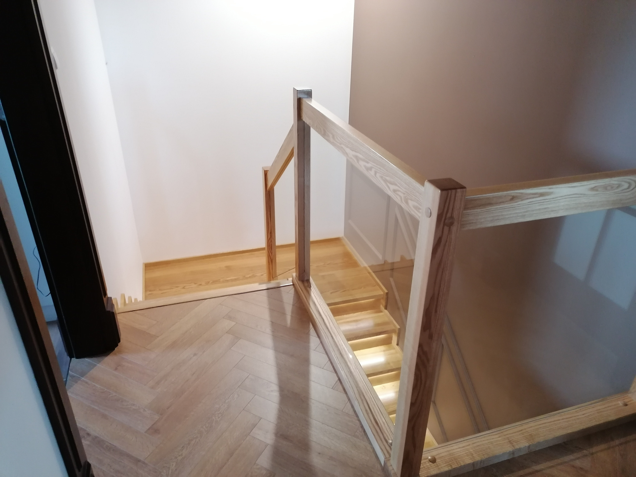 Nowoczesne schody z drewnianymi stopniami i szklaną balustradą, widok z góry na dół, oświetlenie LED pod stopniami.