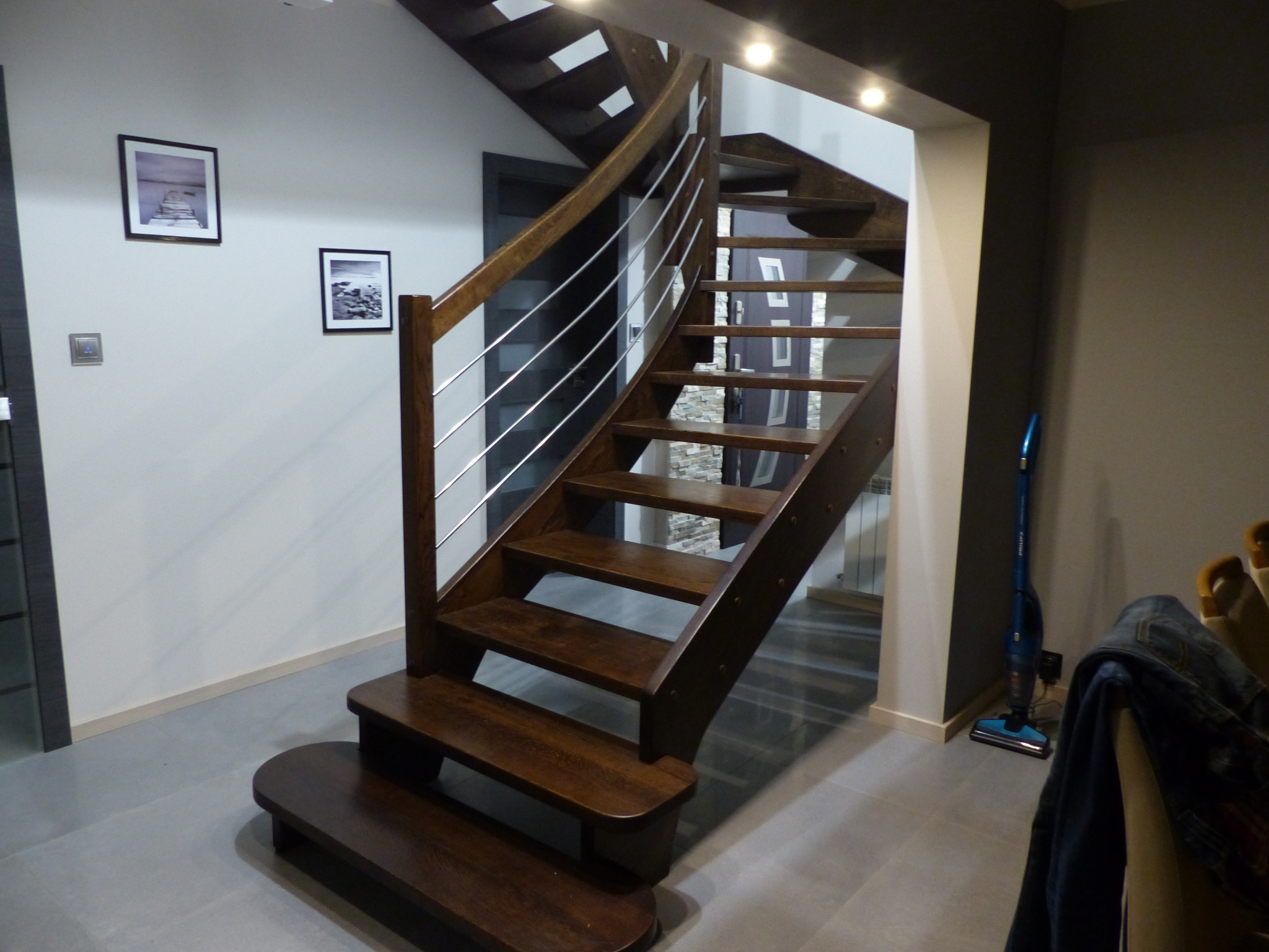 Ciemnobrązowe schody drewniane z metalowymi barierkami, zakręcające w lewo, z oświetleniem punktowym w suficie, na ścianie wiszą dwa obrazy w czarnych ramkach.