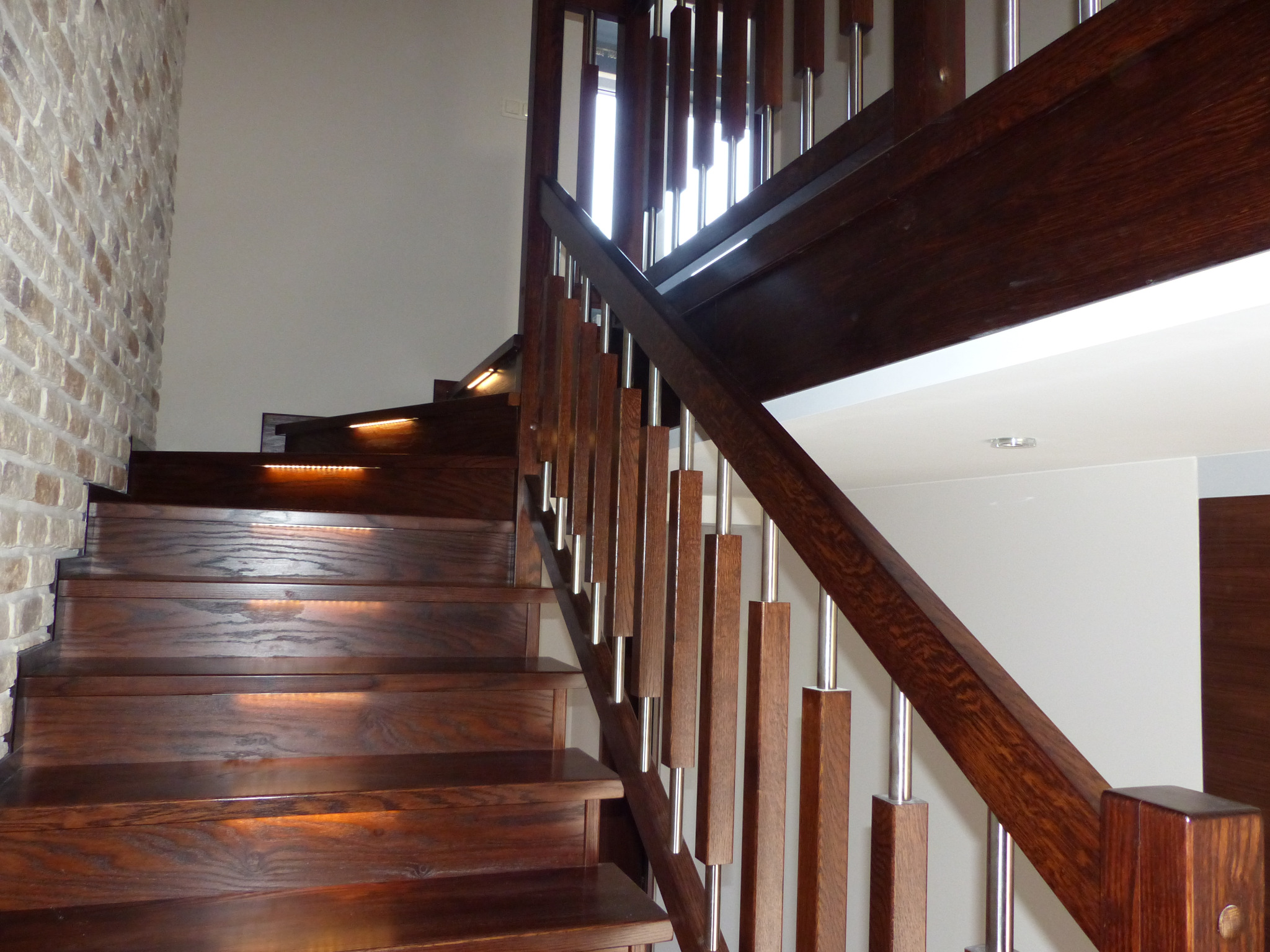 Drewniane schody z ciemnego drewna z nowoczesnymi balustradami z metalowymi prętami, oświetlone od spodu, ściana z dekoracyjnej cegły w tle.