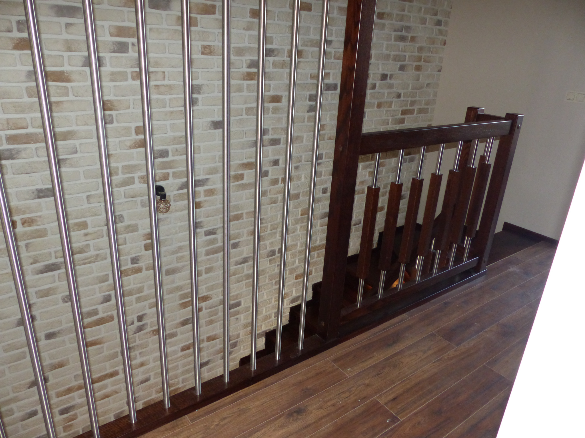 Fragment schodów z drewnianymi stopniami i balustradą z ciemnego drewna oraz metalowymi prętami, ściana wyłożona cegłą dekoracyjną w odcieniach beżu i brązu, podłoga z paneli imitujących drewno.