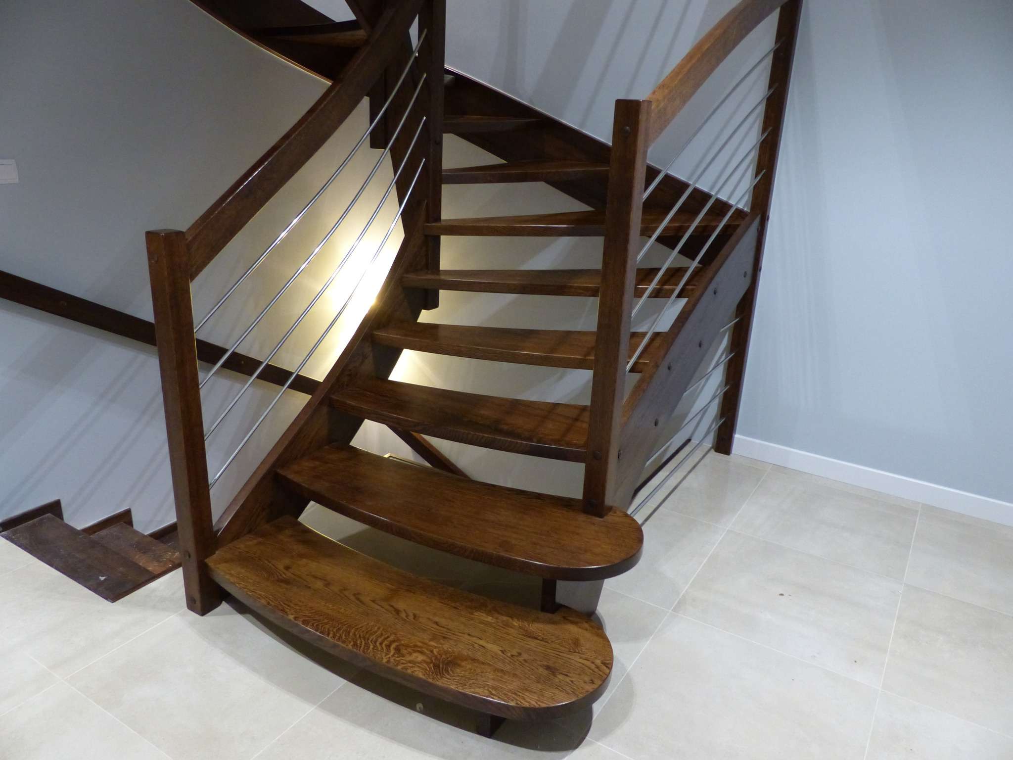 Drewniane schody z metalowymi barierkami, zaokrąglone stopnie, widok z dołu, jasna podłoga z płytek.