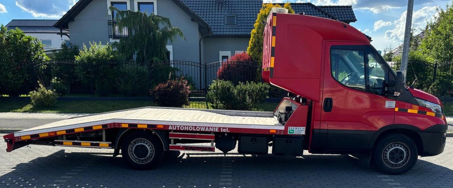 Czerwona laweta na tle domu jednorodzinnego z napisem 'AUTOHOLOWANIE tel.'