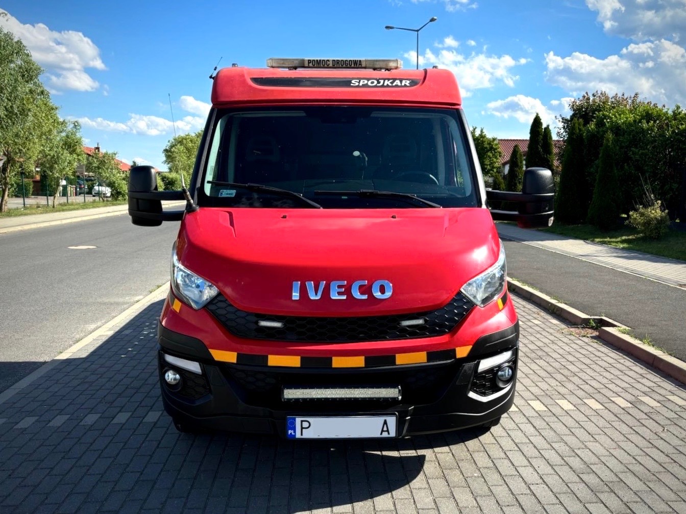 Czerwony pojazd pomocy drogowej marki Iveco, z napisem 'Pomoc Drogowa Spojkar' na górze, zaparkowany na brukowanej drodze w słoneczny dzień.