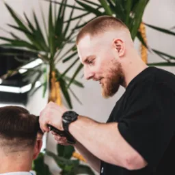 Mężczyzna z brodą w czarnej koszulce strzyże włosy klientowi maszynką, w tle widoczna roślina doniczkowa.