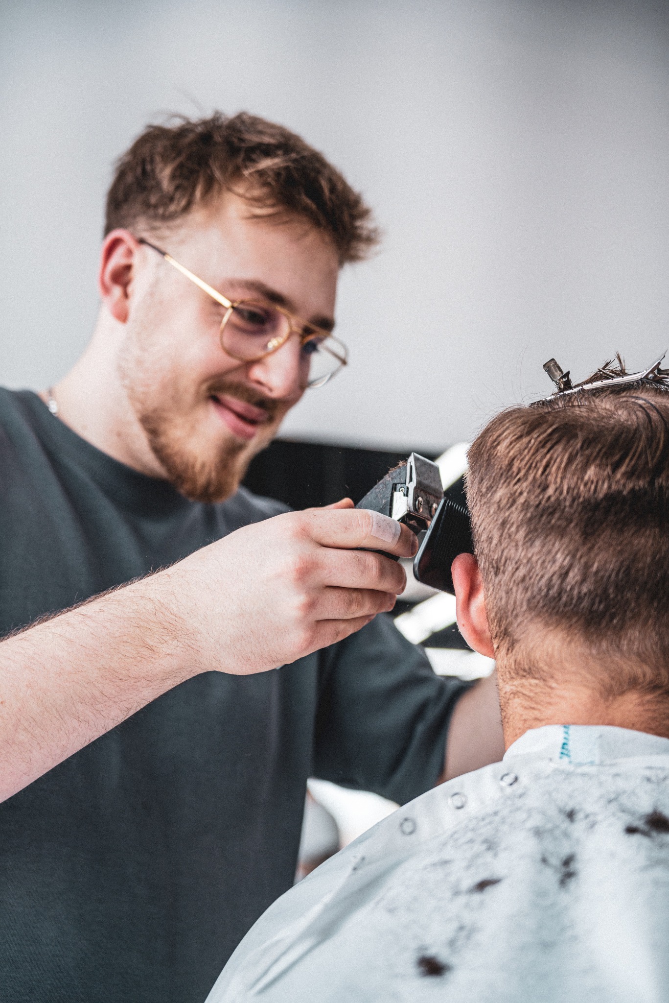 Młody mężczyzna w okularach strzyże włosy klientowi maszynką elektryczną, widoczne klipsy przytrzymujące włosy.