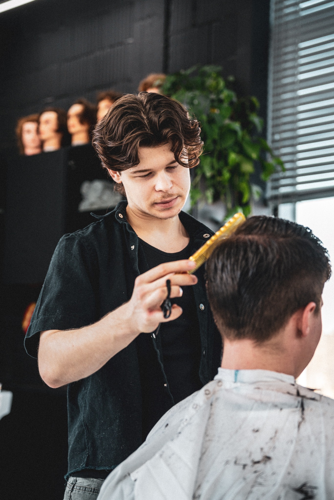 Młody mężczyzna w czarnej koszuli strzyże włosy klientowi za pomocą grzebienia i nożyczek w salonie z dekoracyjną rośliną i manekinami w tle.