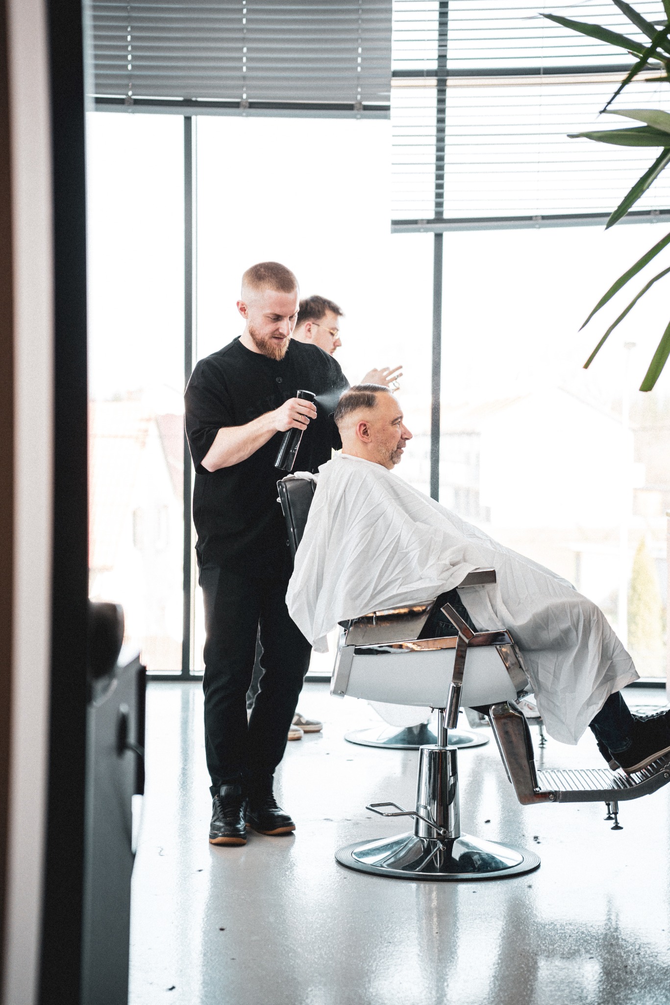 Fryzjer spryskuje włosy klienta wodą przed strzyżeniem w nowoczesnym salonie z dużym oknem i rośliną doniczkową, drugi fryzjer w tle.