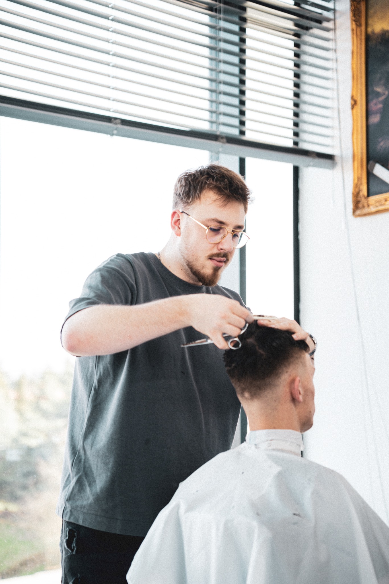 Młody mężczyzna w okularach, z lekkim zarostem, strzyże włosy klientowi przy dużym oknie z żaluzjami, w tle obraz w złotej ramie.