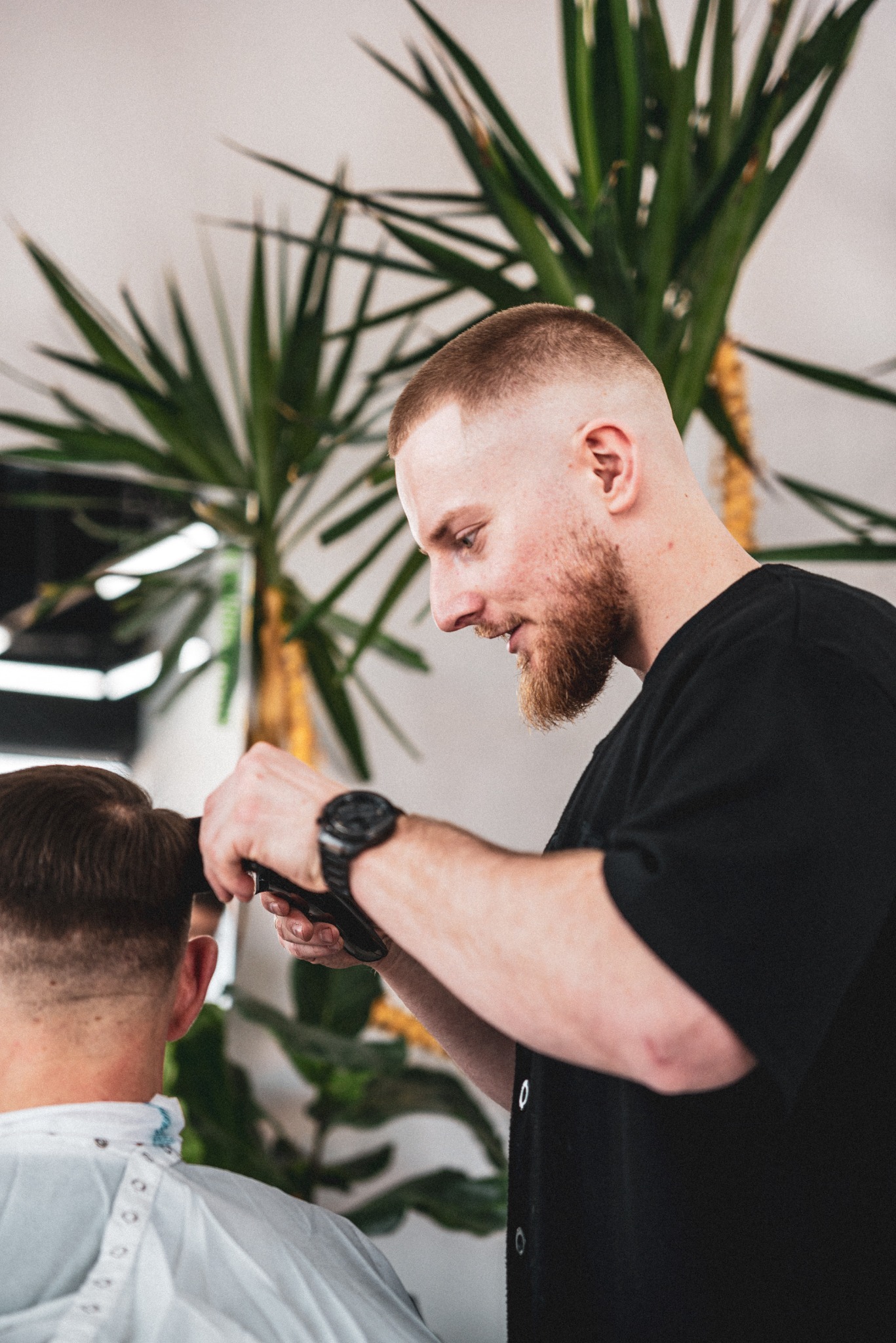 Mężczyzna z brodą w czarnej koszulce strzyże włosy klientowi maszynką, w tle widoczna roślina doniczkowa.