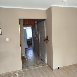 Seweryn Borowczyk - Beżowe ściany w odcieniach ziemi i jasna podłoga z paneli, widok na korytarz z szarą ościeżnicą. Na ścianie włącznik światła i kontakt.