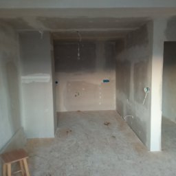 Seweryn Borowczyk - Wnętrze w trakcie szpachlowania: ściany pokryte gładzią, widoczne przewody elektryczne, drewniany stołek w rogu. Etap prac wykończeniowych przed malowaniem.