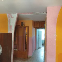 Seweryn Borowczyk - Pokój w trakcie remontu: fragment pomarańczowej ściany z odchodzącą farbą, różowy filar i widok na dalsze pomieszczenia. Widoczny mebel częściowo zakryty folią.