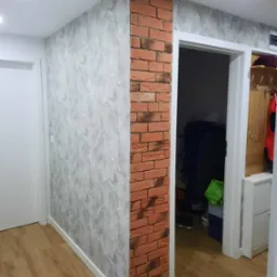 Korytarz po remoncie: ściana z efektem betonu, narożnik wykończony cegłą dekoracyjną. Widoczne białe drzwi i fragment podłogi z paneli. Oświetlenie sufitowe.