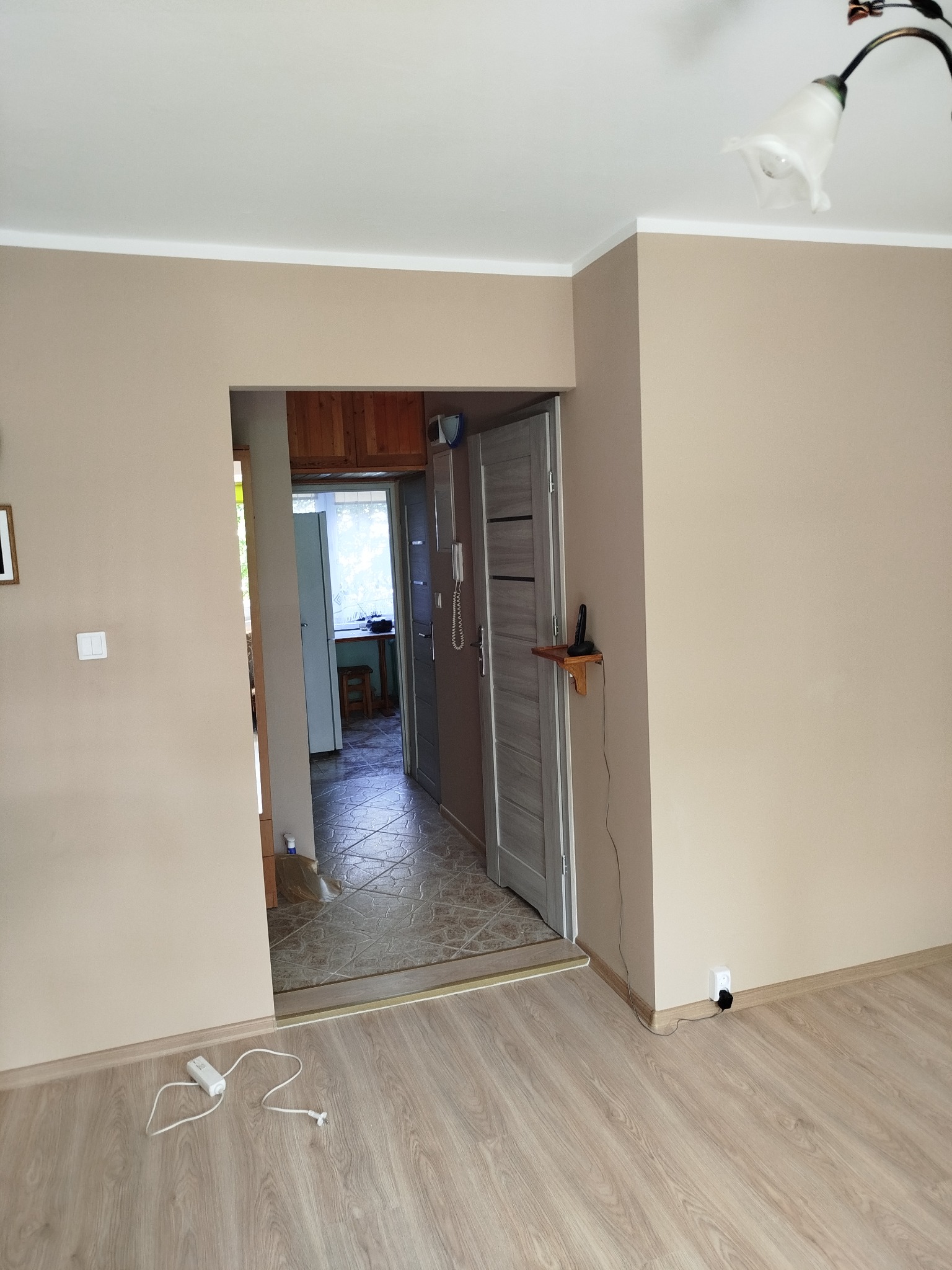 Beżowe ściany w odcieniach ziemi i jasna podłoga z paneli, widok na korytarz z szarą ościeżnicą. Na ścianie włącznik światła i kontakt.
